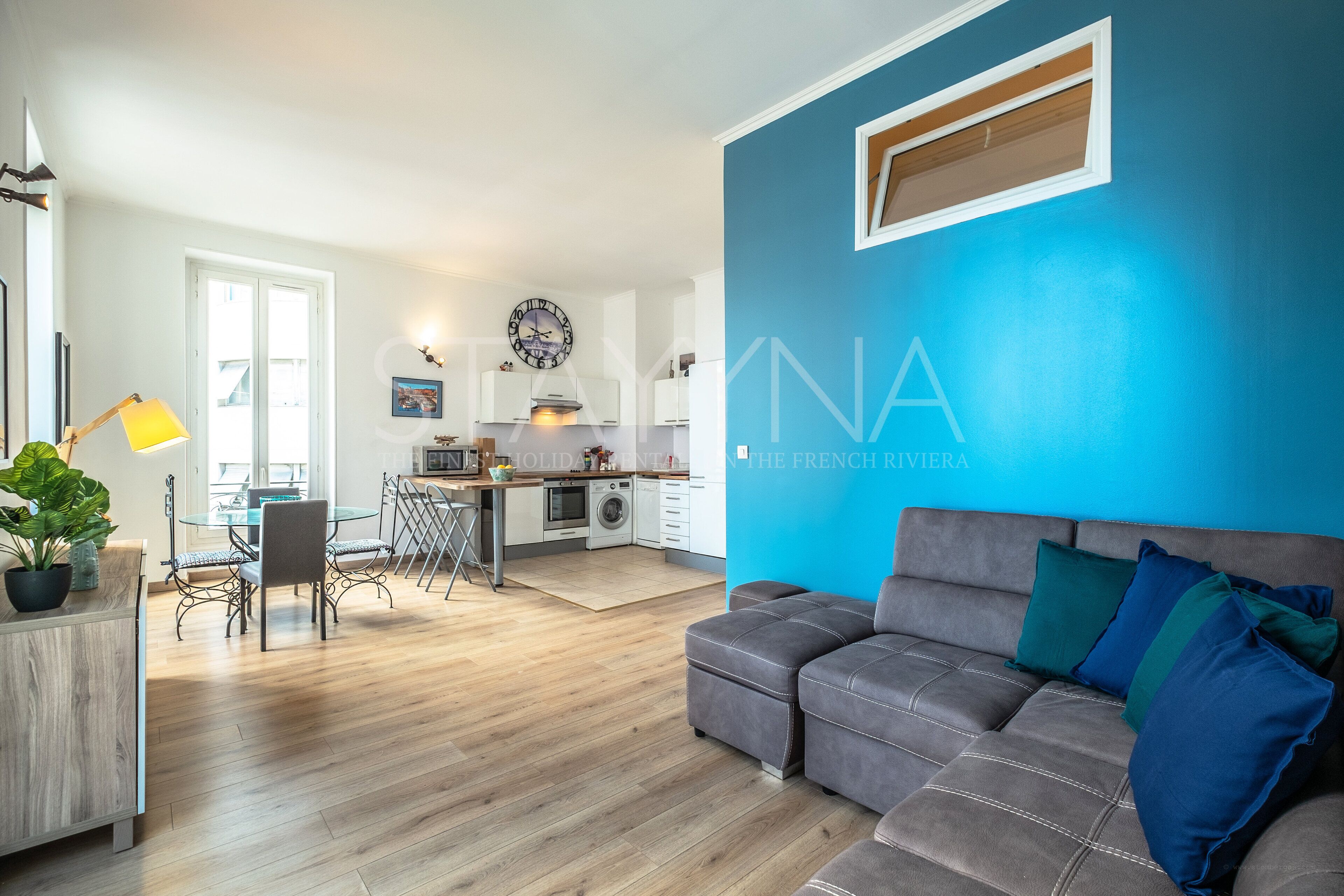 A modern living room with a grey sofa, a dining area with a table and chairs, and a kitchen in the background.