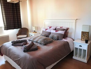 Ein ordentlich gemachtes Bett mit einer braunen Tagesdecke und Kissen, ein Korbstuhl, ein Nachttisch mit Lampe und ein Beistelltisch mit einer Uhr.