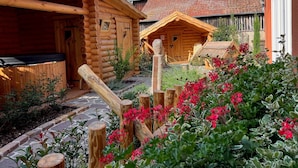 Una cabaña de troncos con un camino de piedra que lleva a una terraza de madera, rodeada por un jardín con flores rojas.