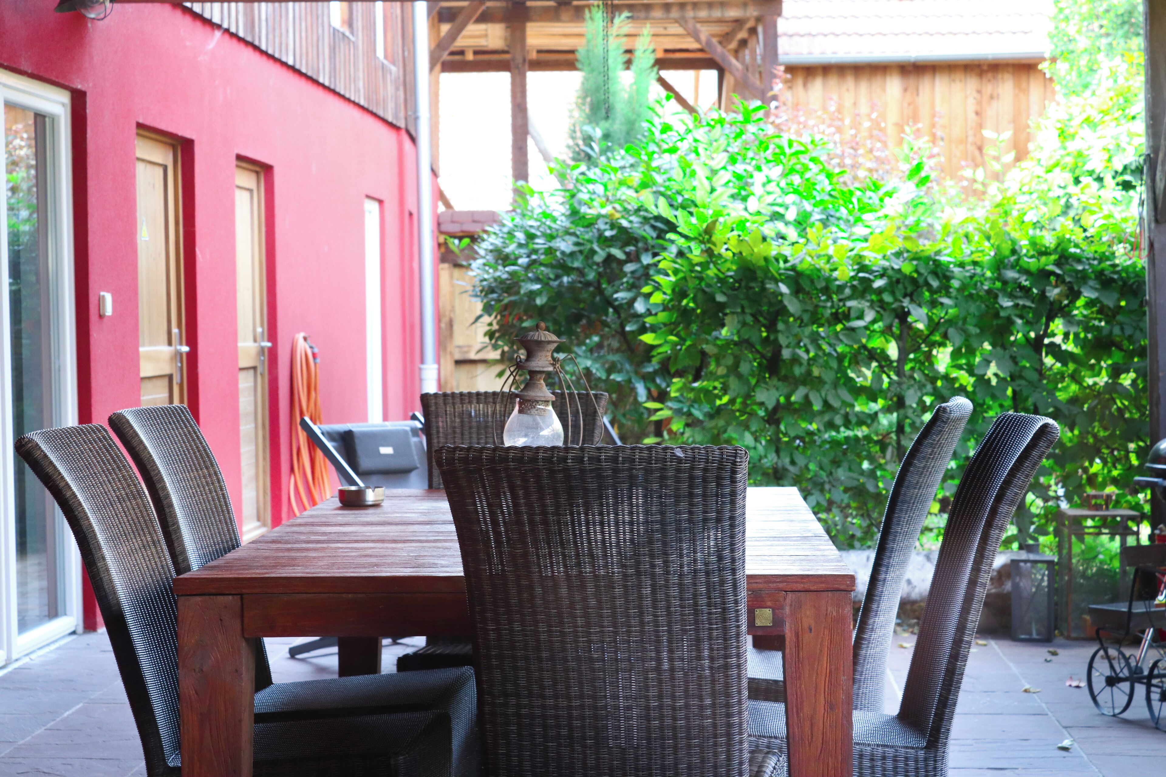 Une terrasse extérieure avec une table en bois et des chaises en osier, entourée d’un bâtiment rouge et de verdure.