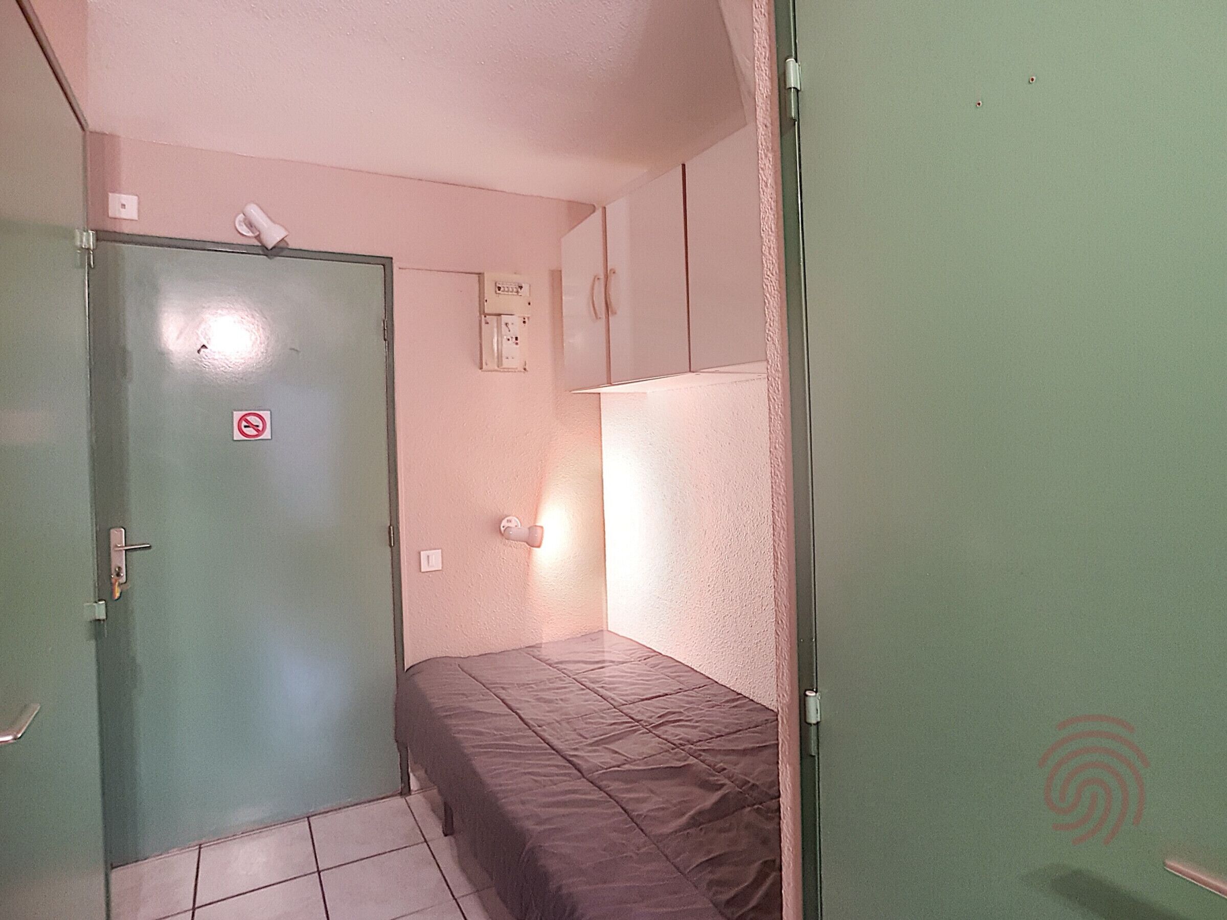 A small, narrow room with a single bed, a door with a no-smoking sign, and a light switch on the wall.