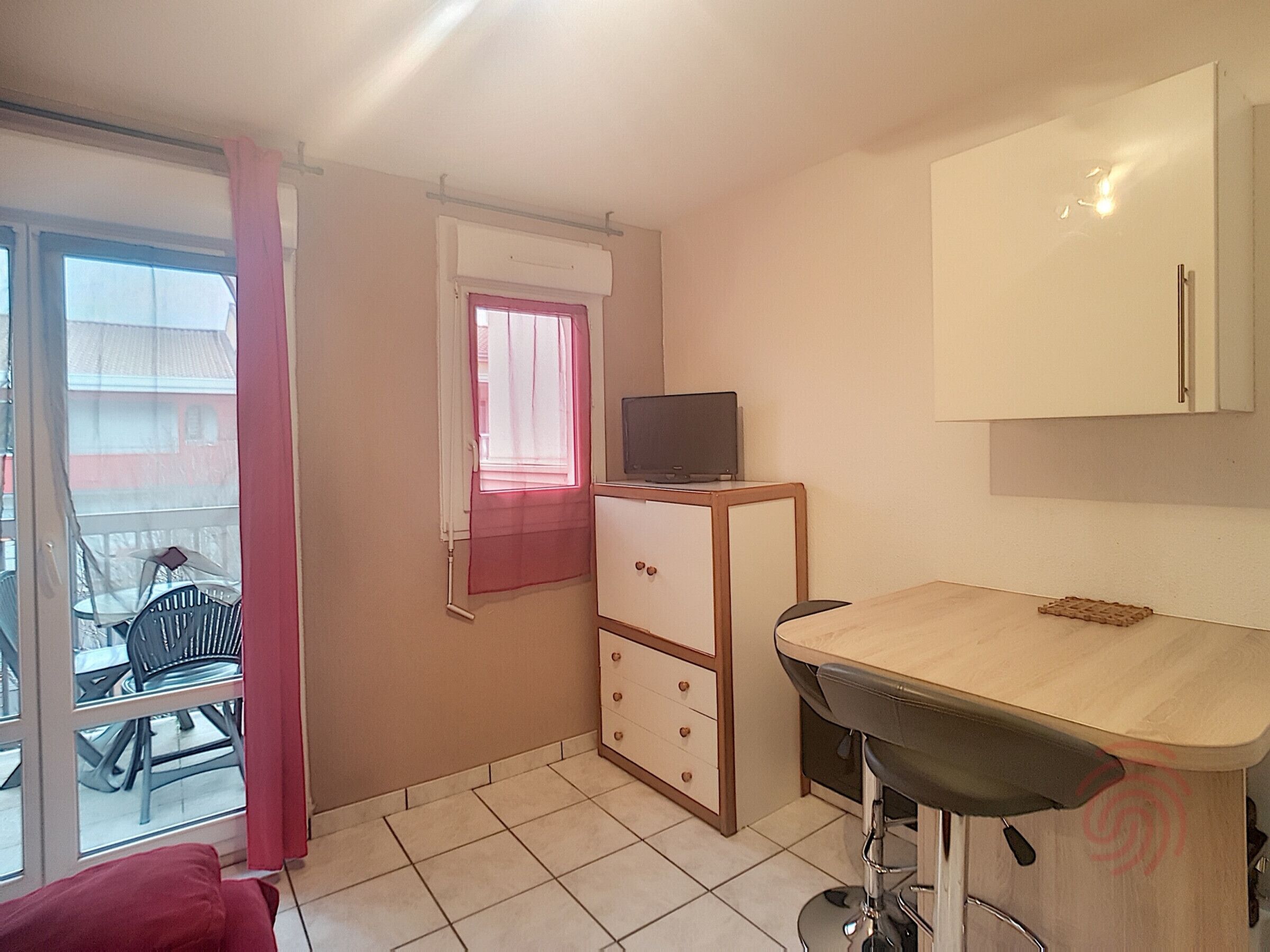 Une petite pièce comprenant un bureau, une chaise, un téléviseur posé sur un meuble, une fenêtre avec un rideau et une vue sur le balcon extérieur.