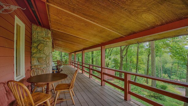 A wooden deck with a stone wall, a table, and chairs, overlooking a forested area.