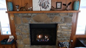 A stone fireplace with a fire, wooden shelves above, and a cozy seating area with a cushioned chair and a small table with magazines.