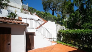 Un bâtiment blanc avec un toit recouvert de tuiles rouges, un escalier menant à l’étage et un jardin clos.