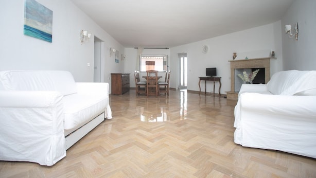 Un salon spacieux avec deux canapés blancs, un parquet en bois disposé en chevrons, une table à manger avec des chaises, une cheminée et un téléviseur.
