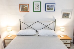 Un lit bien fait, avec du linge de lit blanc et deux lampes de chevet, une table de chevet en bois avec un tiroir, et trois tableaux encadrés accrochés au mur.