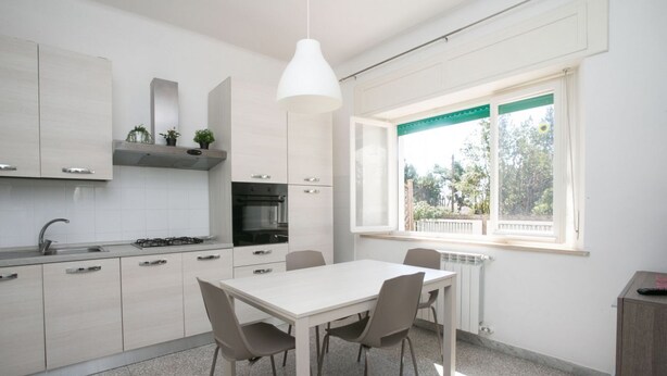 A modern kitchen with white cabinets, a dining table with chairs, a window with a view of greenery, and a hanging light fixture. - N050 - Numana, three-room apartment in the center with porch and parking space (Numana)