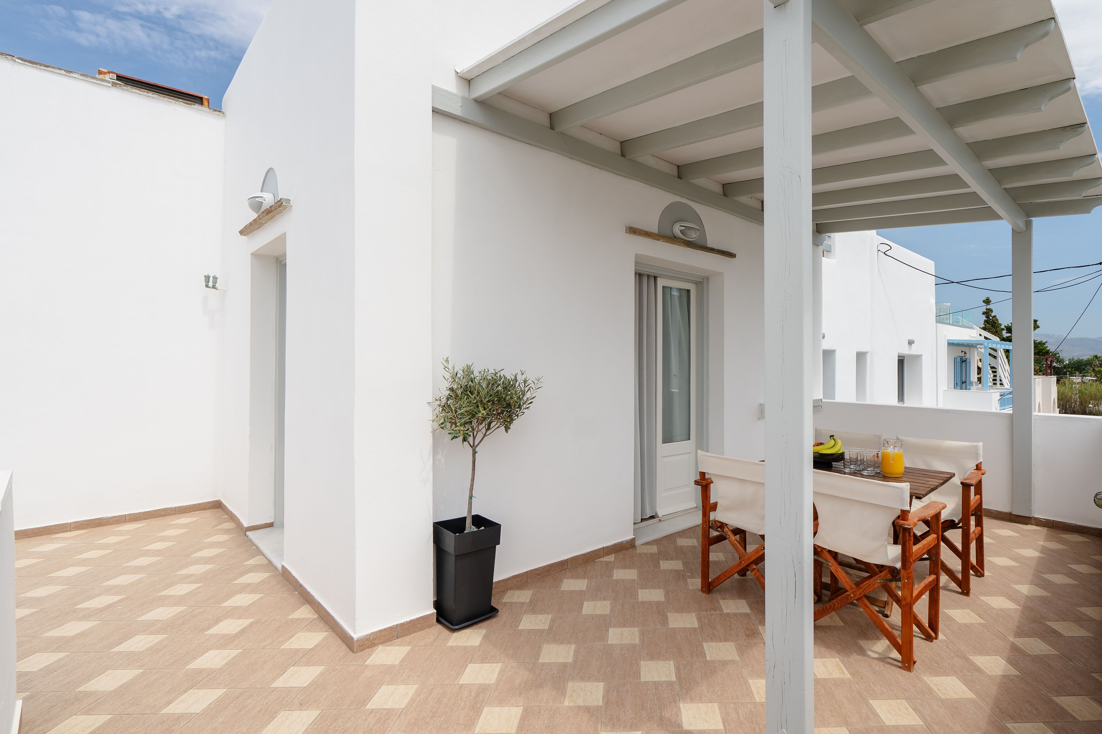 Fachada blanca y limpia de un edificio con un patio de baldosas, una pequeña mesa con sillas y una planta en maceta.