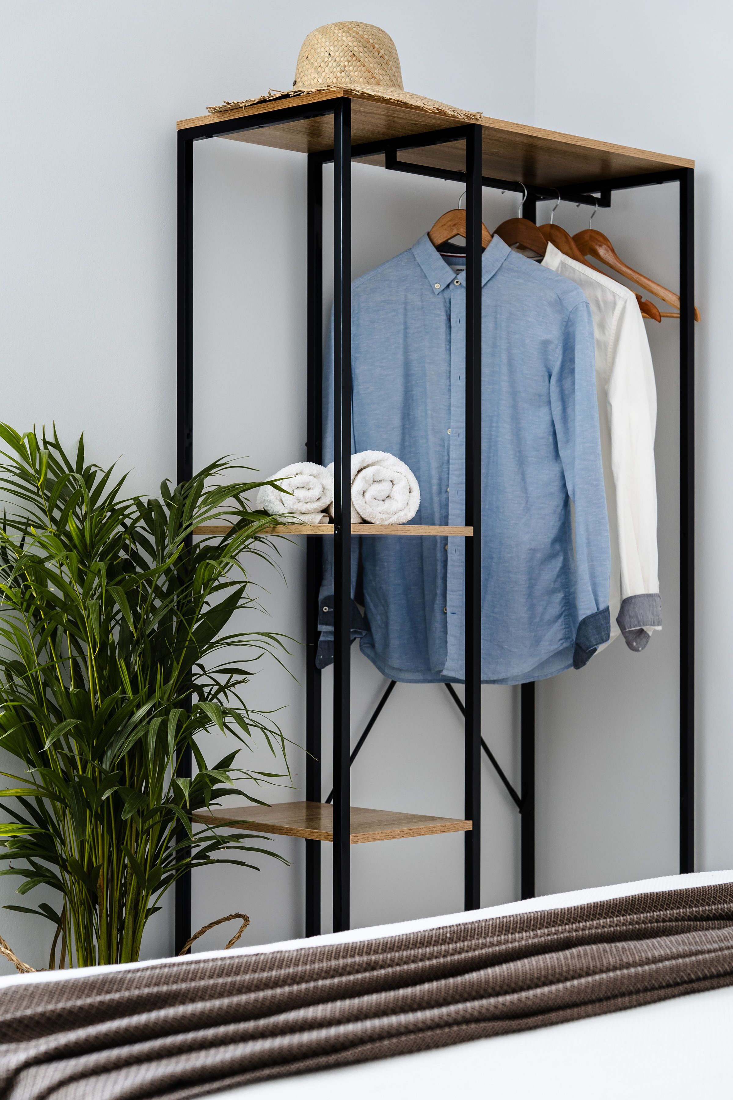 A room with a bed, a plant, a clothing rack with folded clothes, and a hat on top.