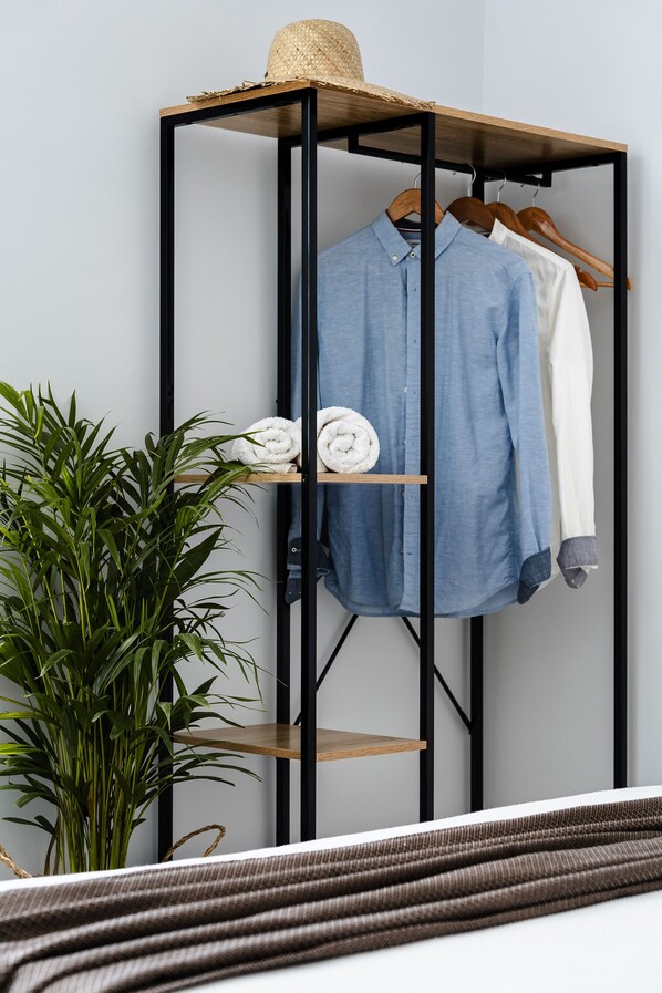 Quarto com cama, uma planta, um cabideiro com roupas dobradas e um chapéu em cima.