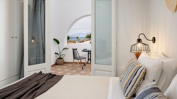 A bedroom with a bed, a bedside table, a lamp, a window with curtains, a view of the outside, and a small balcony with a table and chairs. - Erato Inn (Naxos)