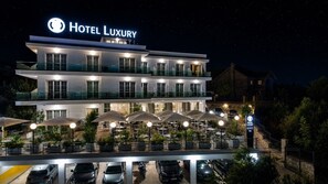 A multi-story hotel with a prominent 'Hotel Luxury' sign, outdoor seating with umbrellas, and a parking area. - Hotel Luxury (Ksamil)