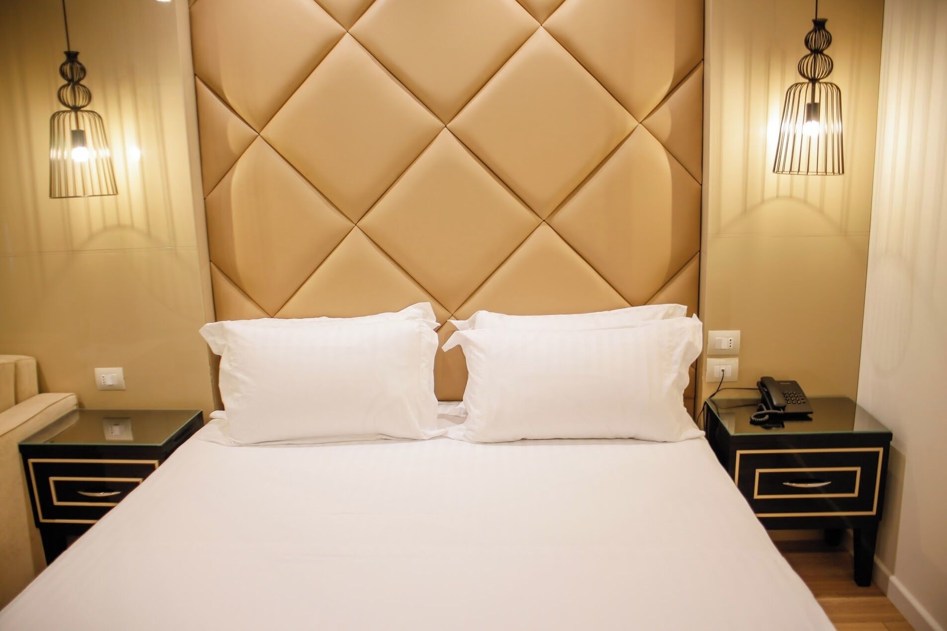 A hotel room with a tufted headboard, two bedside tables with lamps, a bed with white linens, and a telephone on one of the tables.