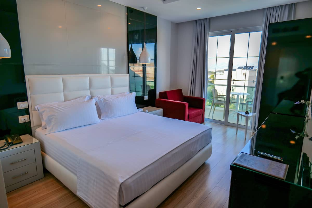 A modern hotel room with a large bed, a red chair, a small table, and a large window with a view of buildings.