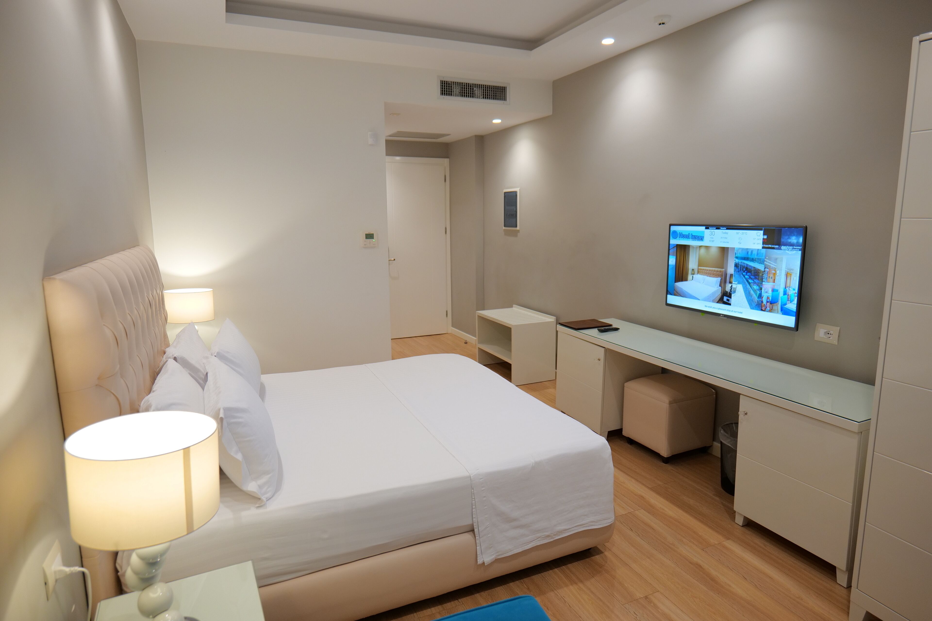 A modern hotel room with a bed, bedside lamp, TV, desk, and storage cabinet.