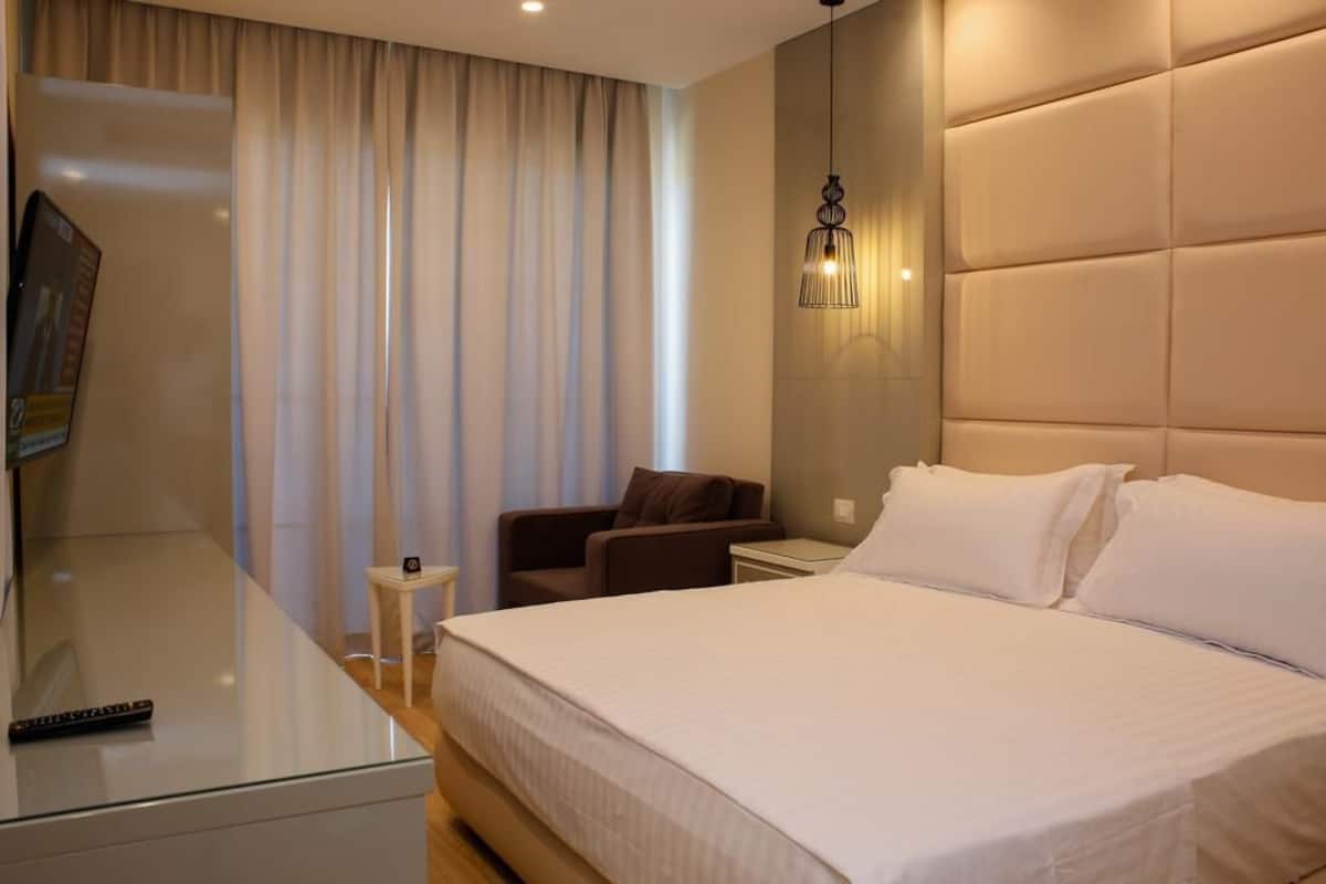 A modern hotel room with a large bed, a small seating area, a TV, and a wall-mounted light fixture.
