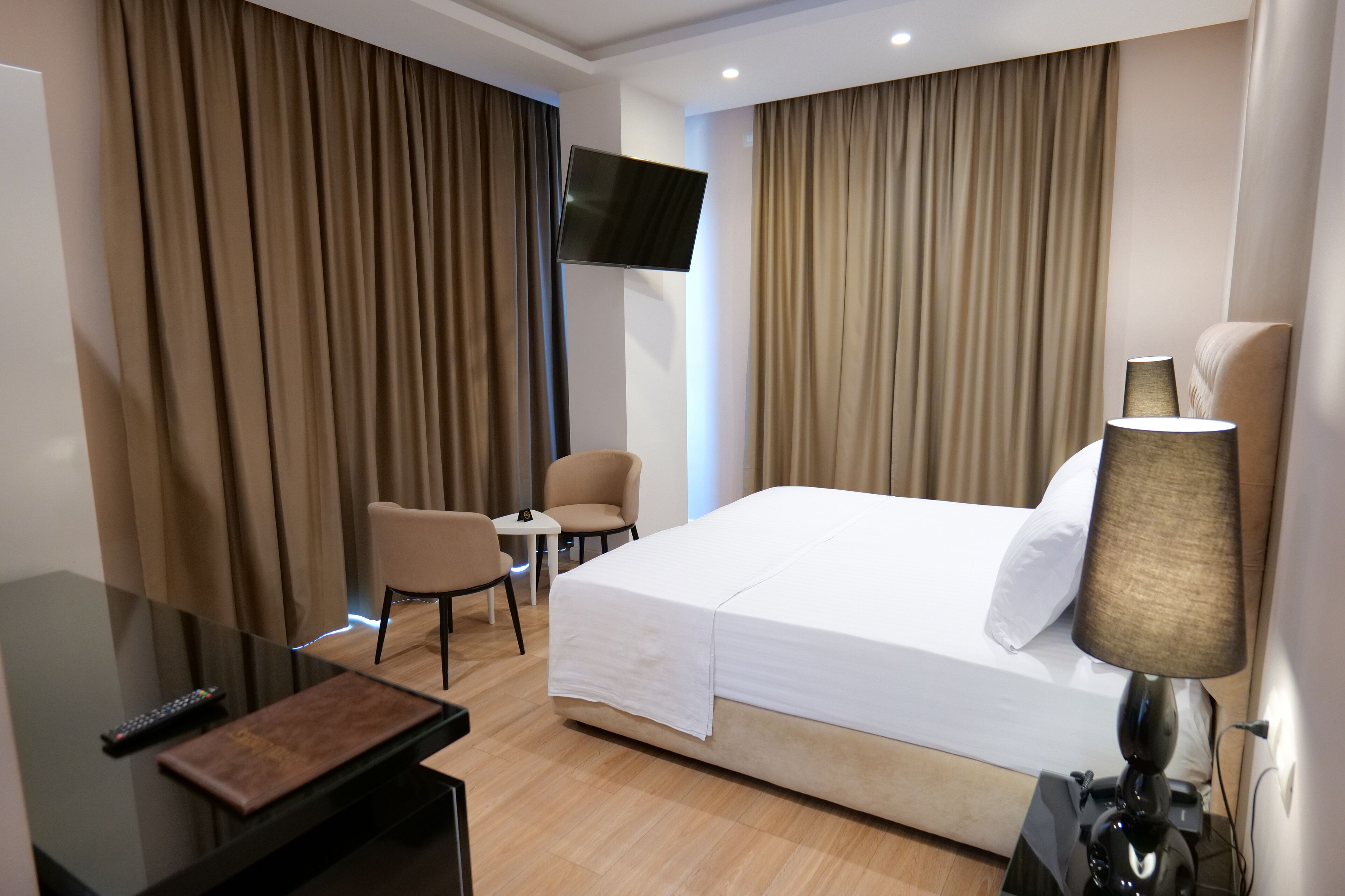 A hotel room with a large bed, a TV mounted on the wall, a desk with a lamp, and two chairs.