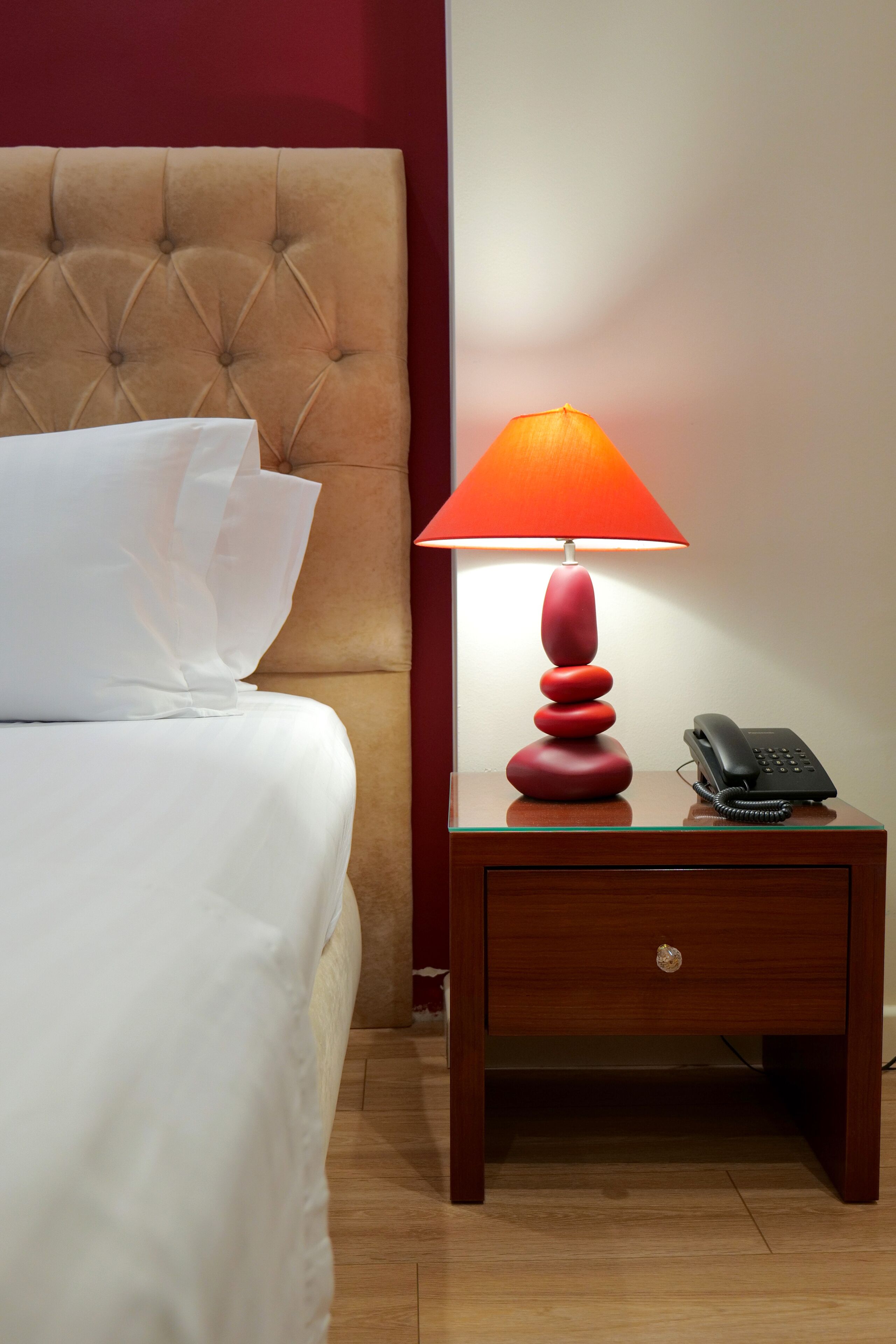 A neatly made bed with white linens and pillows, a red lamp on a wooden nightstand, and a black telephone.