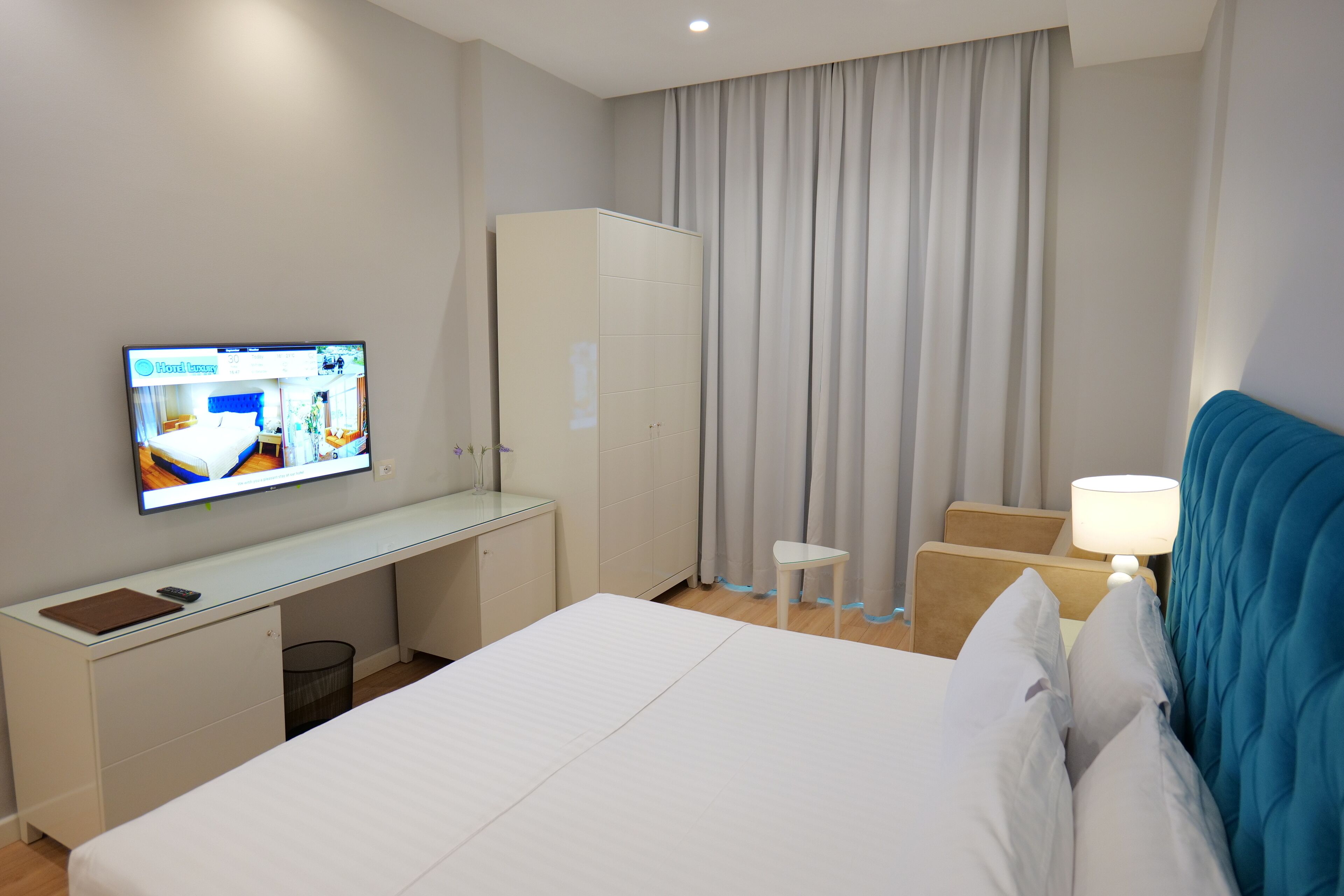 Una habitación de hotel con cama, escritorio, televisor, armario, silla y mesita de noche con lámpara.