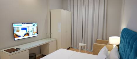 Una habitación de hotel con cama, escritorio, televisor, armario, silla y mesita de noche con lámpara.