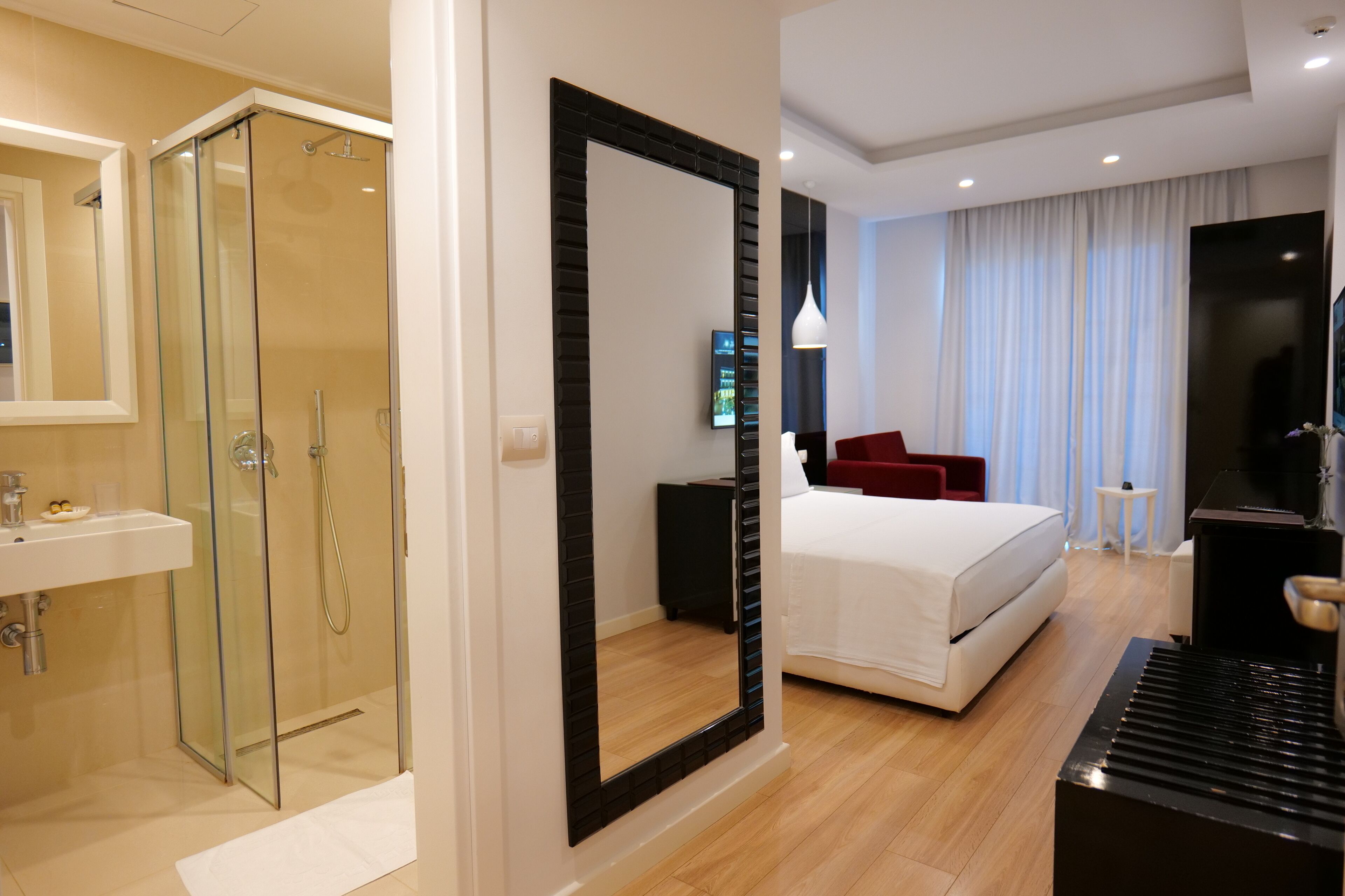 A modern hotel room with a glass-enclosed shower, a large mirror, a bed with white linens, a red chair, and a television.