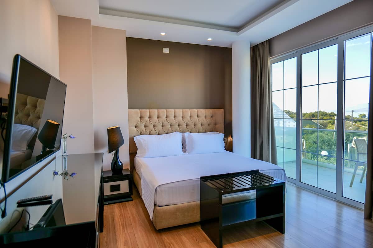 A modern hotel room with a large bed, a desk with a computer, a TV, and a balcony with a view.