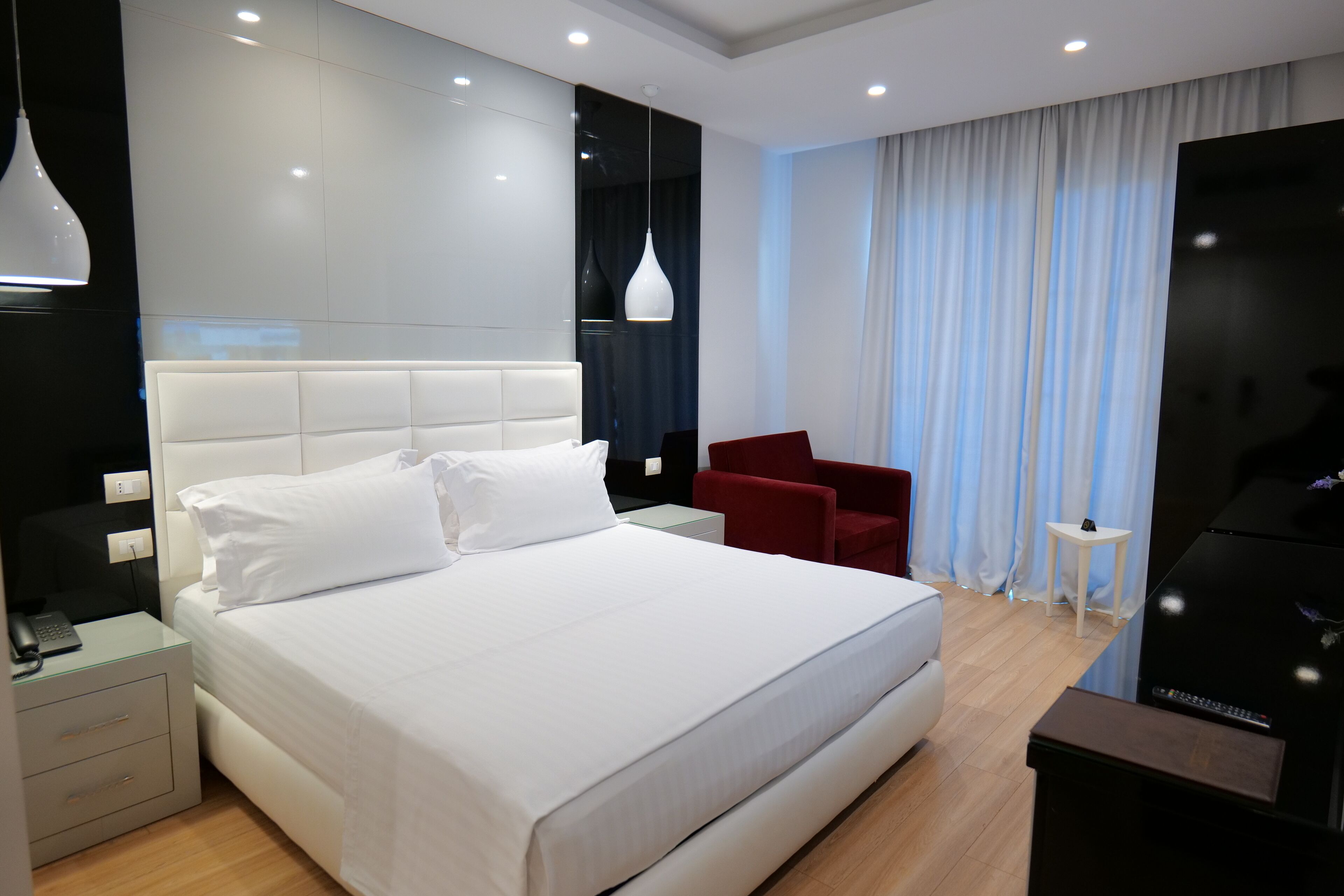 Ein modernes Hotelzimmer mit einem großen Bett, einem Schreibtisch mit Lampe, einem roten Sessel, einem kleinen Tisch und einem Fernseher.