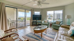 A spacious living room with a large flat-screen TV, comfortable seating, a ceiling fan, and a view of the ocean through large windows. - CRC 2204 - Second Floor Ocean, Pool View Condo (St Augustine)