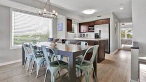 A modern kitchen with a central island, a dining area with six chairs, and a large window with blinds. - Oceanfront Townhome with Amazing Ocean Views (Wrightsville Beach)