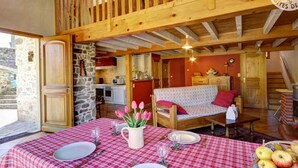 A cozy, rustic room with a wooden balcony, stone walls, a dining table set with a checkered tablecloth, and a fruit basket.