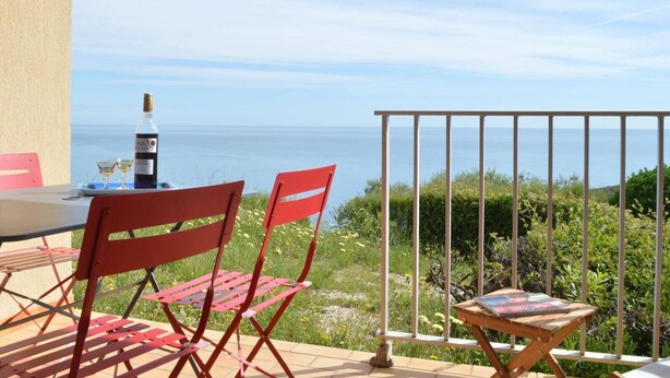 A terrace with a table, two red chairs, a bottle of wine, and a view of the sea.