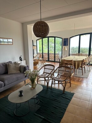 Un amplio salón con sofá, una mesa redonda, sillas de mimbre y una lámpara colgante.