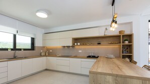 Una cocina moderna con armarios blancos, encimera de madera y una isla integrada. Se observan estantes con adornos y una lámpara colgante.
