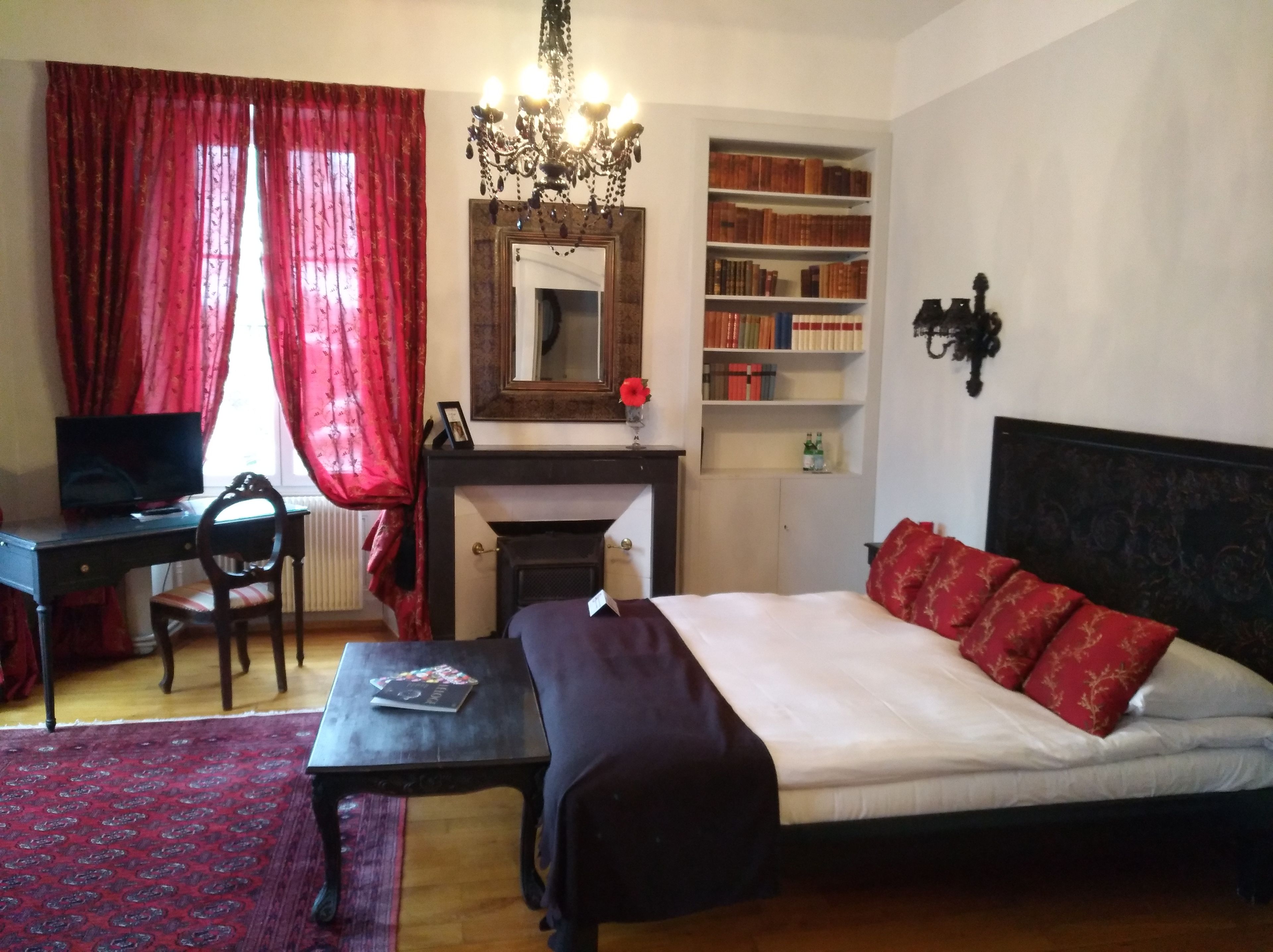 Un dormitorio con cama, escritorio, silla, estantería, chimenea y araña.
