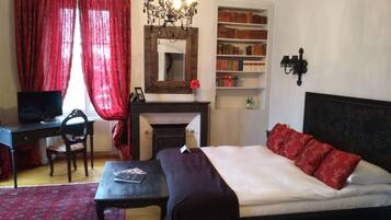 Un dormitorio con cama, escritorio, silla, estantería, chimenea y araña.