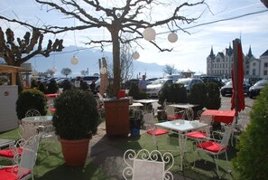 An outdoor seating area with white metal chairs and tables, surrounded by potted plants and a large tree.