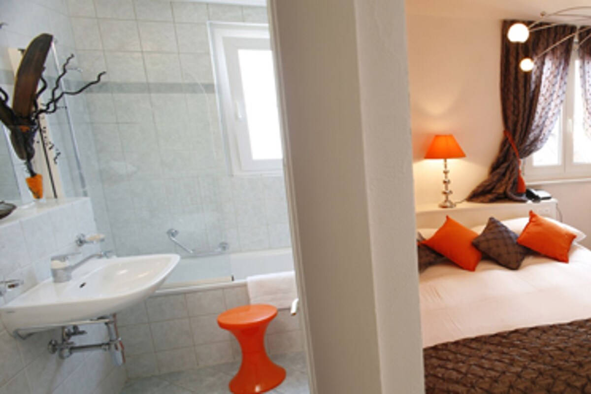 A hotel room with a bed, a desk, a chair, a glass table, a red stool, a white sink, and a bathtub.