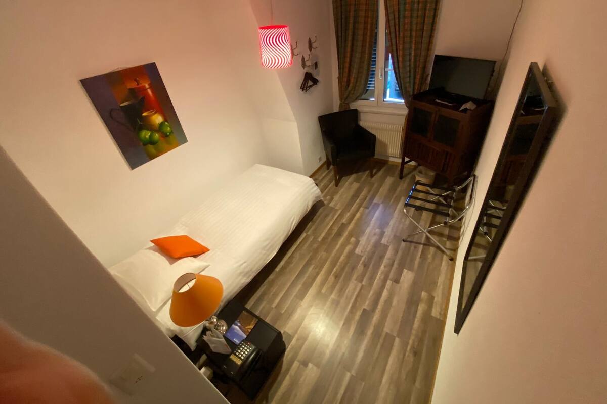 Habitación de hotel con cama, escritorio con lámpara, una silla, televisor y ventana con cortinas.