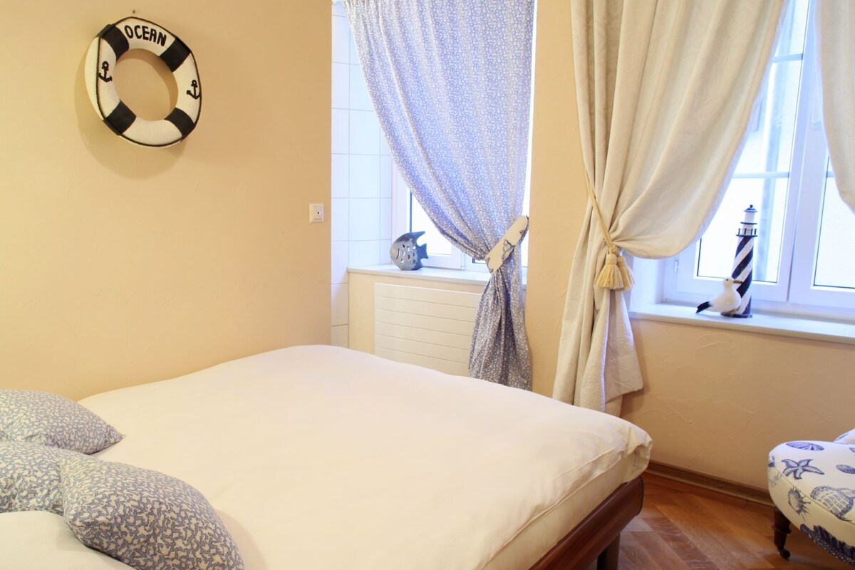 A bedroom with a bed, pillows, a life preserver decoration, a window with curtains, and a chair with a nautical theme.