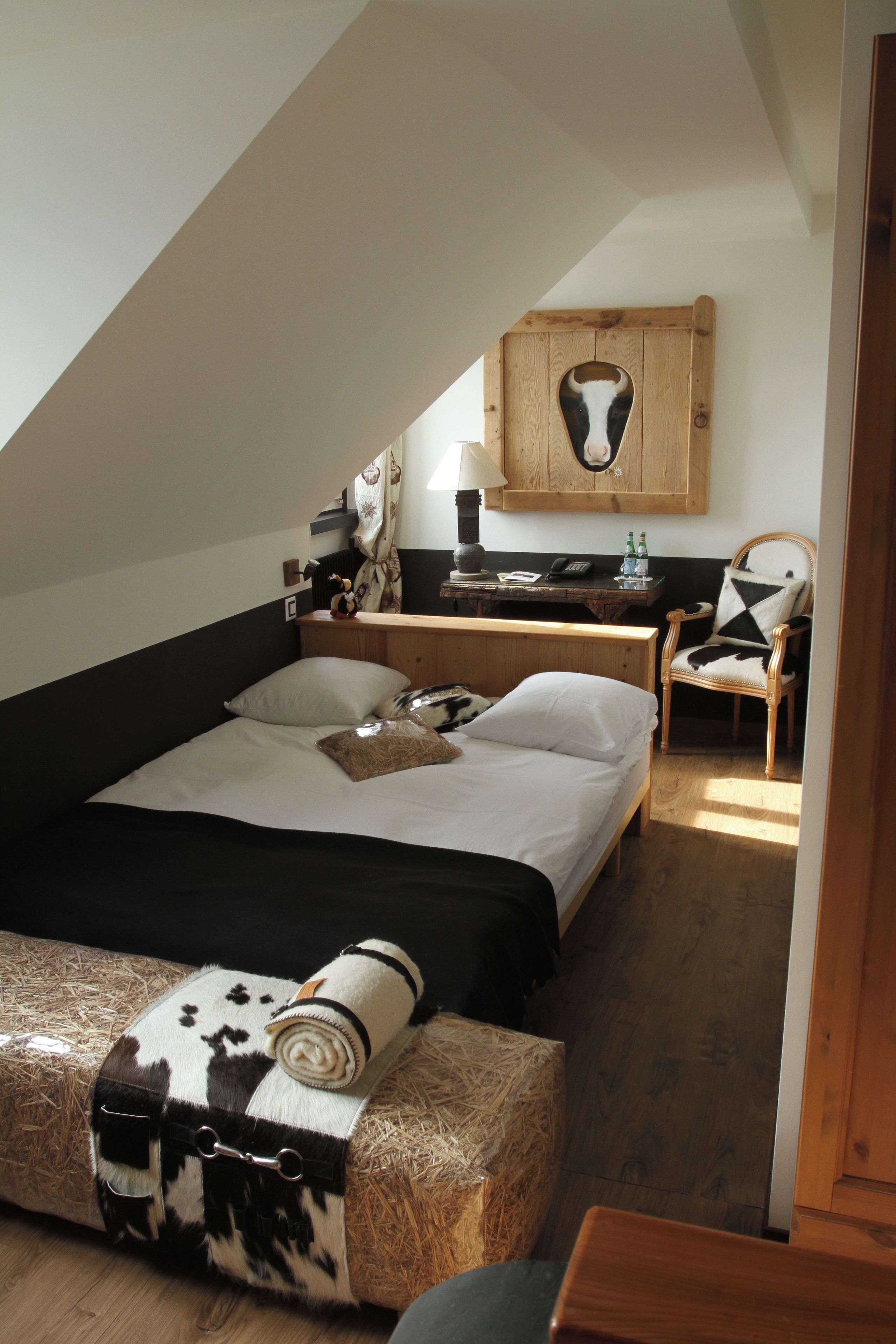 A bedroom with a sloped ceiling, a bed with white and black bedding, a wooden nightstand, a chair with a cow print cushion, and a wall-mounted wooden headboard with a cow head cutout.