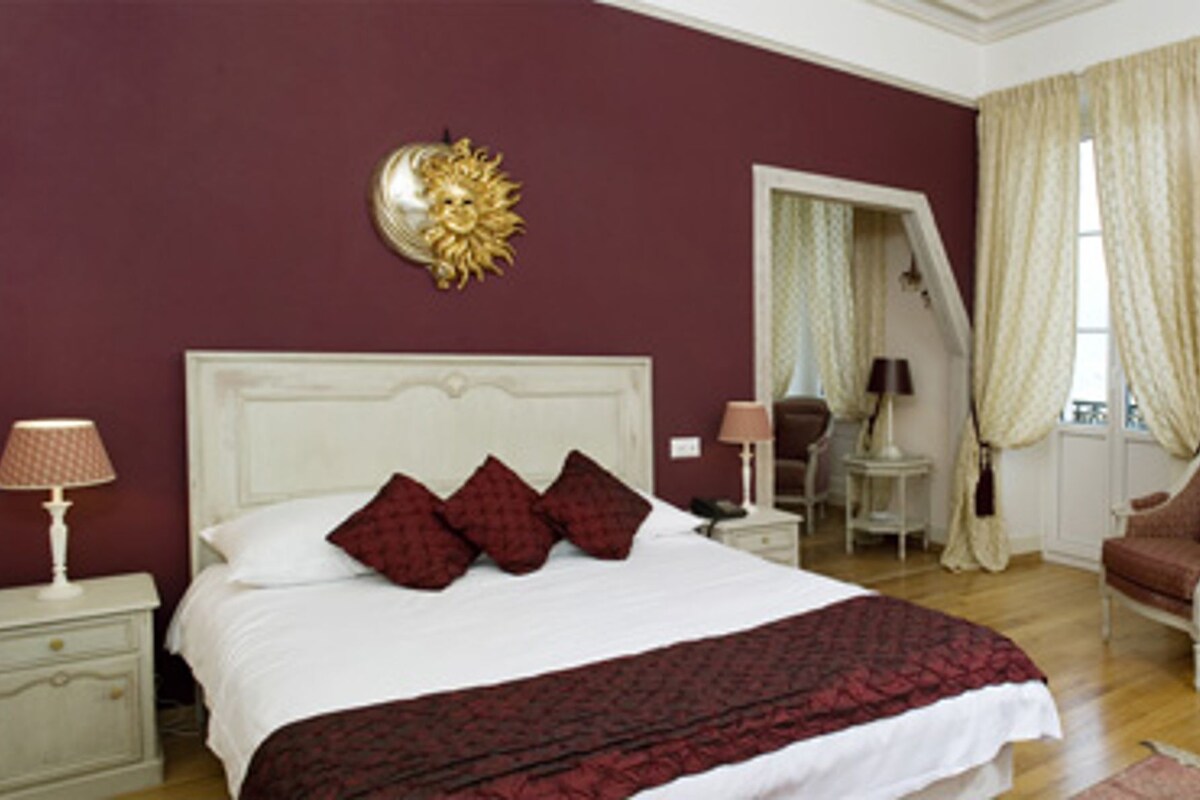 A bedroom with a large bed, a nightstand with a lamp, a chair, a desk, and a window with curtains.