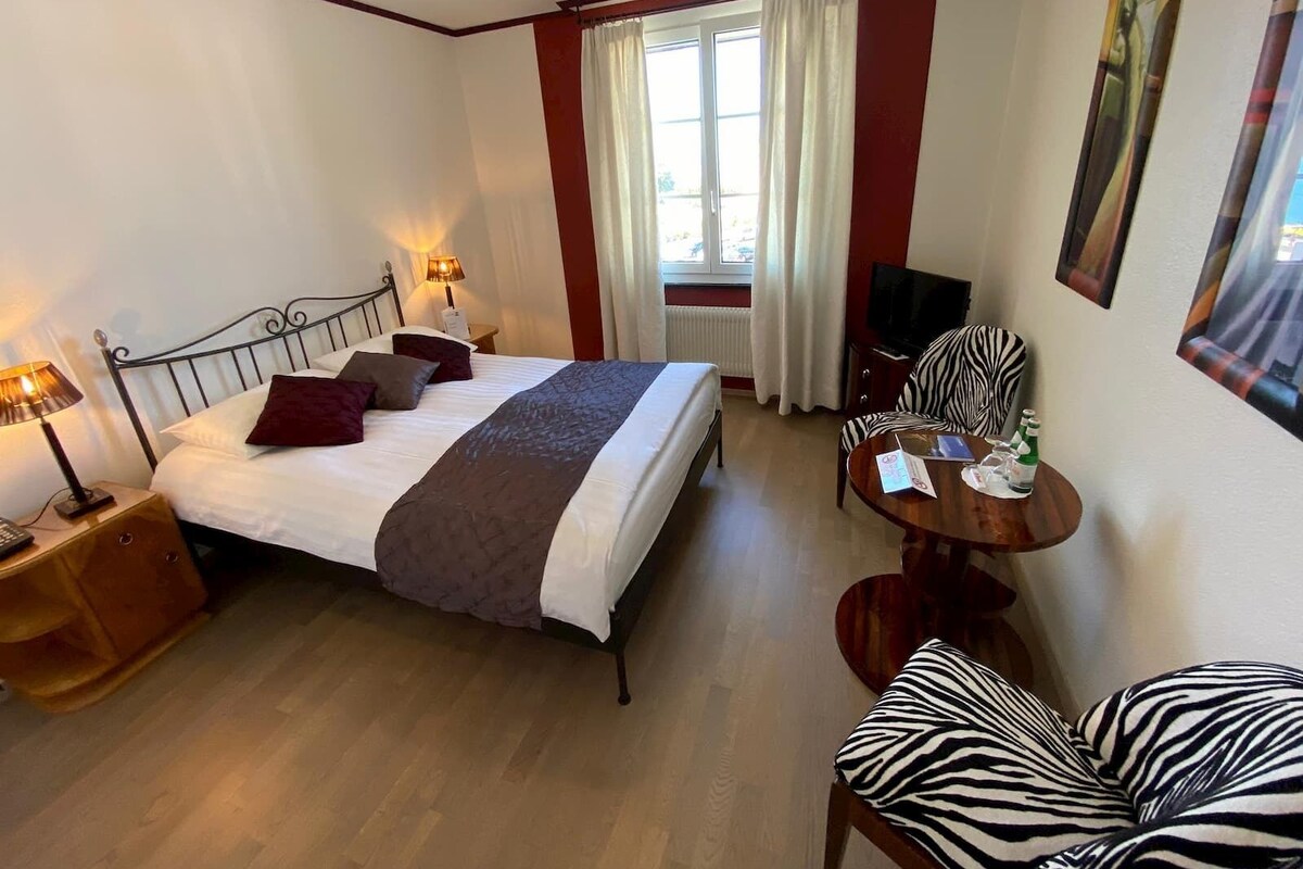A hotel room with a bed, bedside lamps, a TV, a zebra-patterned chair, a small table, and a window with curtains.