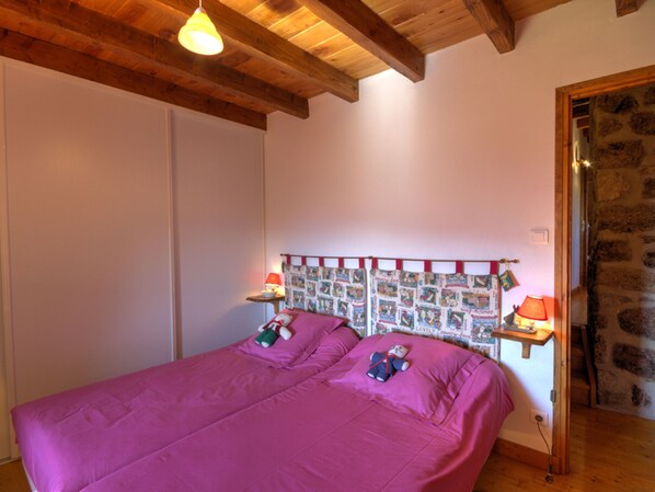 A bedroom with a bed, wooden beams, a quilted headboard, two bedside lamps, and a stone wall.