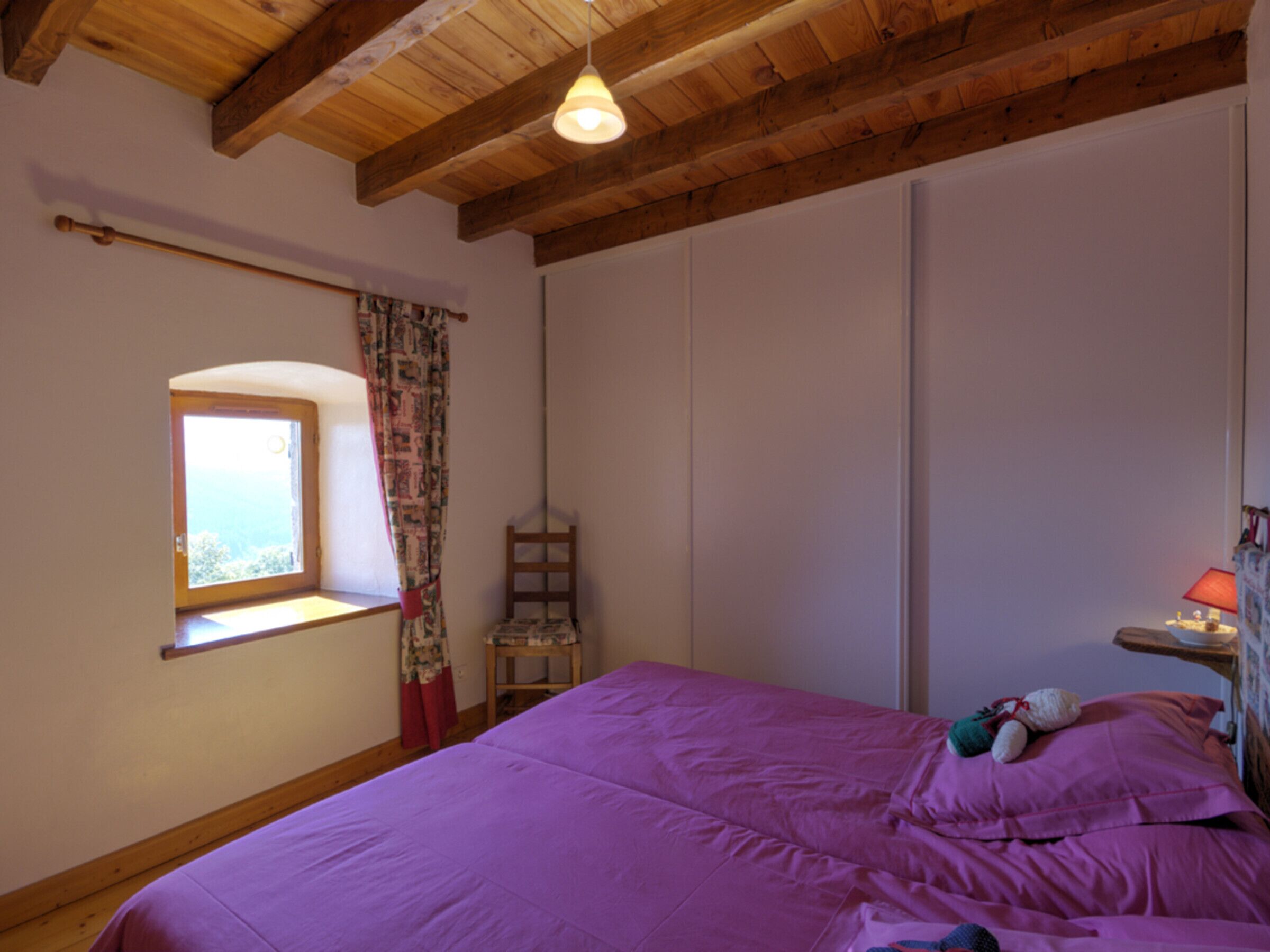 A bedroom with a wooden ceiling, a window with curtains, a bed with a purple bedspread, and a nightstand with a lamp.