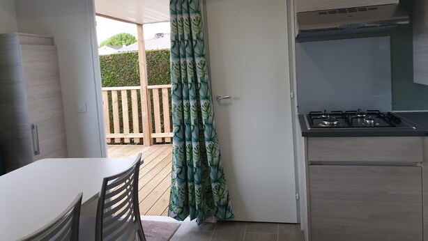 Una cocina compacta con estufa a gas, una mesa de comedor con sillas y un acceso corredizo a un espacio exterior con terraza de madera y seto.