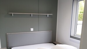 Una cama cuidadosamente arreglada con sábanas y almohadas blancas, un cabecero gris y una ventana con vistas a zonas verdes.