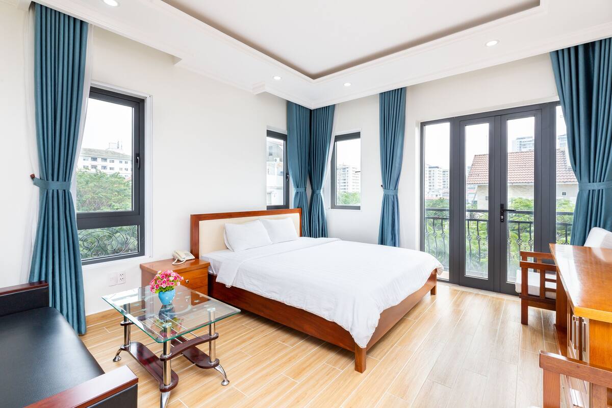 A hotel room with a bed, a glass table with a vase, a wooden desk, and a balcony with curtains.
