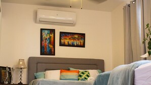 A bedroom with a bed, a nightstand, an air conditioner, and two paintings on the wall.