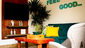 A cozy living room with a wooden coffee table, a green sofa with a yellow pillow, and a wall-mounted TV. A large plant and a welcome sign are also visible.
